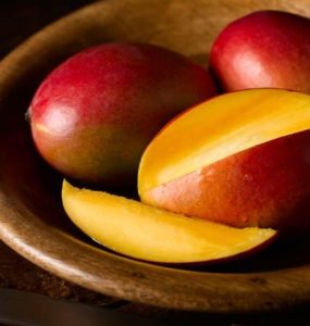 Eine in Scheiben geschnittene Mango, dahinter zwei ganze, rote Mangos