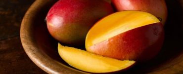 Eine in Scheiben geschnittene Mango, dahinter zwei ganze, rote Mangos