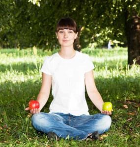 Frau in Jeans und weißem Tshirt sitz im Schneidersitz auf grünem Rasen mit einem roten Apfel in der rechten und einem grünen Apfel in der anderen Hand.