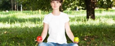 Frau in Jeans und weißem Tshirt sitz im Schneidersitz auf grünem Rasen mit einem roten Apfel in der rechten und einem grünen Apfel in der anderen Hand.