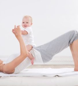 Junge Frau mit dunklen Haaren und weißem T Shirt liegt auf dem Rücken auf einem weißen Handtuch und hebt die Hüfte an. Ein lachendes Baby sitzt auf ihrem Schoß.