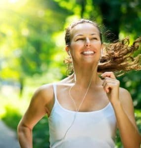 Junge Frau mit weißem Trägertop joggt durch den Wald, lacht und hört Musik.