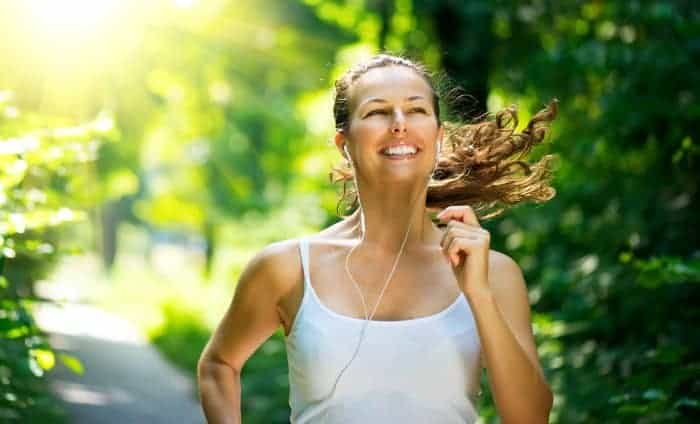 Junge Frau mit weißem Trägertop joggt durch den Wald, lacht und hört Musik.