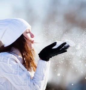 Frau mit weißer Mütze und Schal pustet Schnee aus ihren Händen