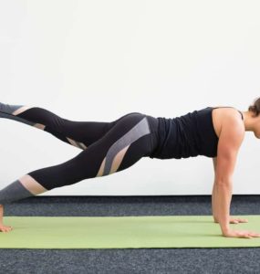 Junge Frau in Liegestützposition auf einer hellgrünen Trainingsmatte macht Pilates Leg Pull Front