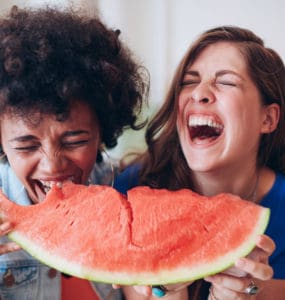 Zwei junge Frauen lachen und essen zusammen ein großes Stück Wassermelone