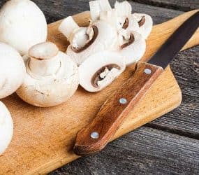 Weiße Champignons und ein Messer auf einem Holzbrett, zum Teil am Stück, zum Teil geschnitten