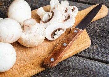 Weiße Champignons und ein Messer auf einem Holzbrett, zum Teil am Stück, zum Teil geschnitten