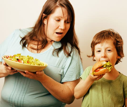 Mutter mut Salatteller in der Hand neben ihrem Sohn, der einen Burger isst