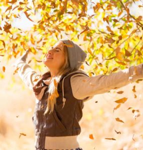 Junge Frau mit Jacke und Mütze streckt die Arme nach oben und ist glücklich. Herbst Laub fliegt im Hintergrund durch die Luft