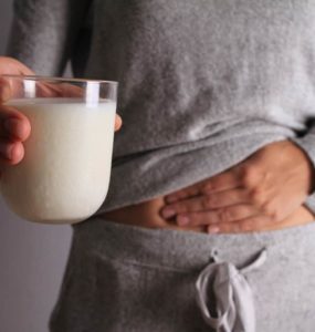 Frau mit Milchglas in der Hand fasst sich an den Bauch, hat Blähungen durch Laktoseintoleranz