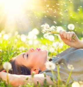 Junge Frau genießt den Moment liegt auf einer Wiese und pustet gegen eine Pusteblume.